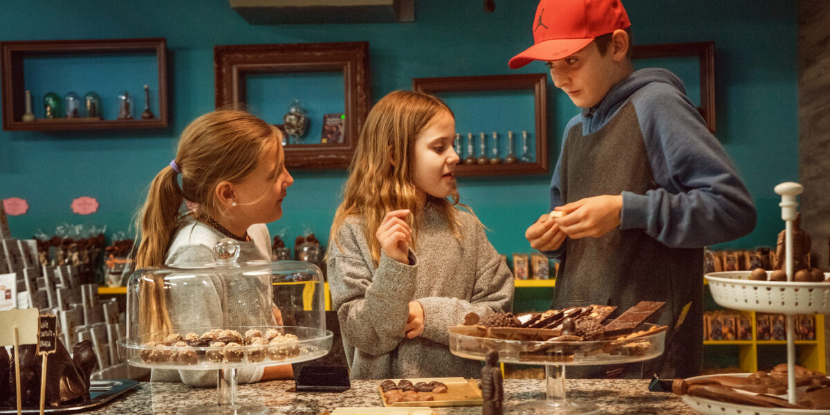 Drei Kinder stehen an der Theke eines Schokoladenladens, betrachten verschiedene Schokoladensorten, die auf Tabletts und Ständern ausgestellt sind, und unterhalten sich darüber.