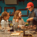 Drei Kinder stehen an der Theke eines Schokoladenladens, betrachten verschiedene Schokoladensorten, die auf Tabletts und Ständern ausgestellt sind, und unterhalten sich darüber.