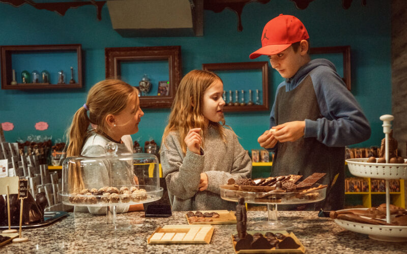 Drei Kinder stehen an der Theke eines Schokoladenladens, unterhalten sich und wählen Pralinen aus verschiedenen Auslagen aus.
