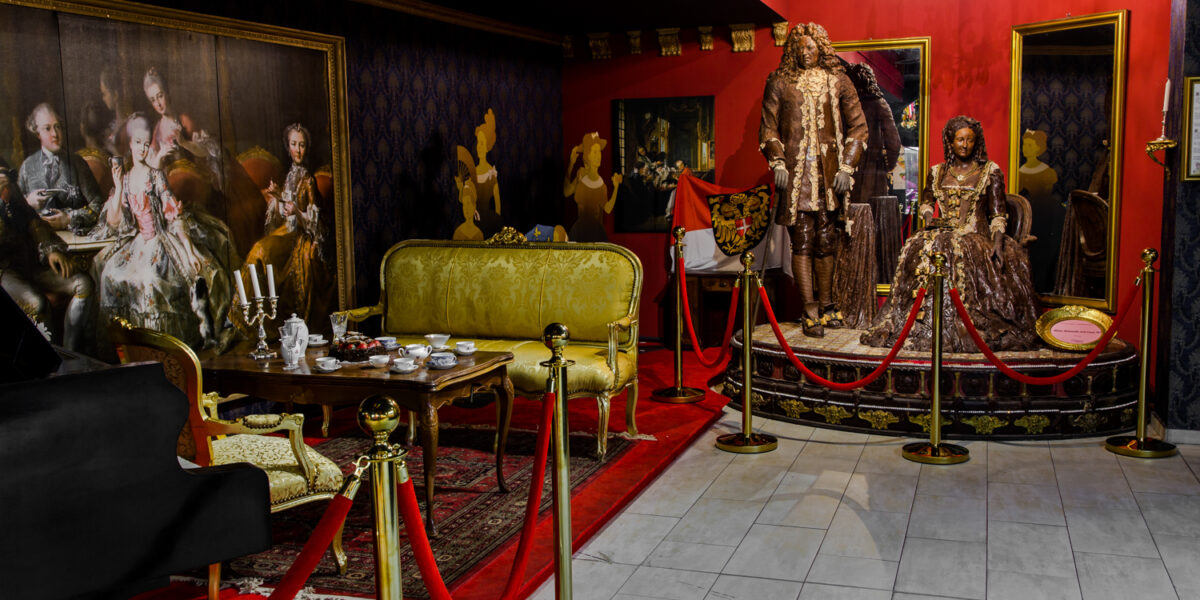 Eine Museumsausstellung mit Schaufensterpuppen in kunstvollen historischen Kostümen, antiken Möbeln, einem Kronleuchter und klassischen Gemälden, getrennt durch rote Seile auf goldenen Ständern.