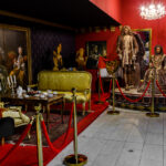 Eine Museumsausstellung mit Schaufensterpuppen in kunstvollen historischen Kostümen, antiken Möbeln, einem Kronleuchter und klassischen Gemälden, getrennt durch rote Seile auf goldenen Ständern.