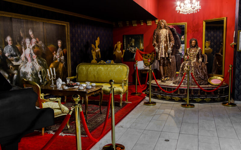 Eine Museumsausstellung mit Schaufensterpuppen in kunstvollen historischen Kostümen, antiken Möbeln, einem Kronleuchter und klassischen Gemälden, getrennt durch rote Seile auf goldenen Ständern.