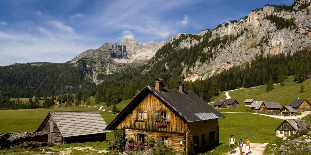 Eine Gruppe von Menschen geht auf einem Weg vorbei an Holzhäusern in einem grasbewachsenen Alpental, mit Bergen und Kiefernwäldern im Hintergrund unter blauem Himmel.