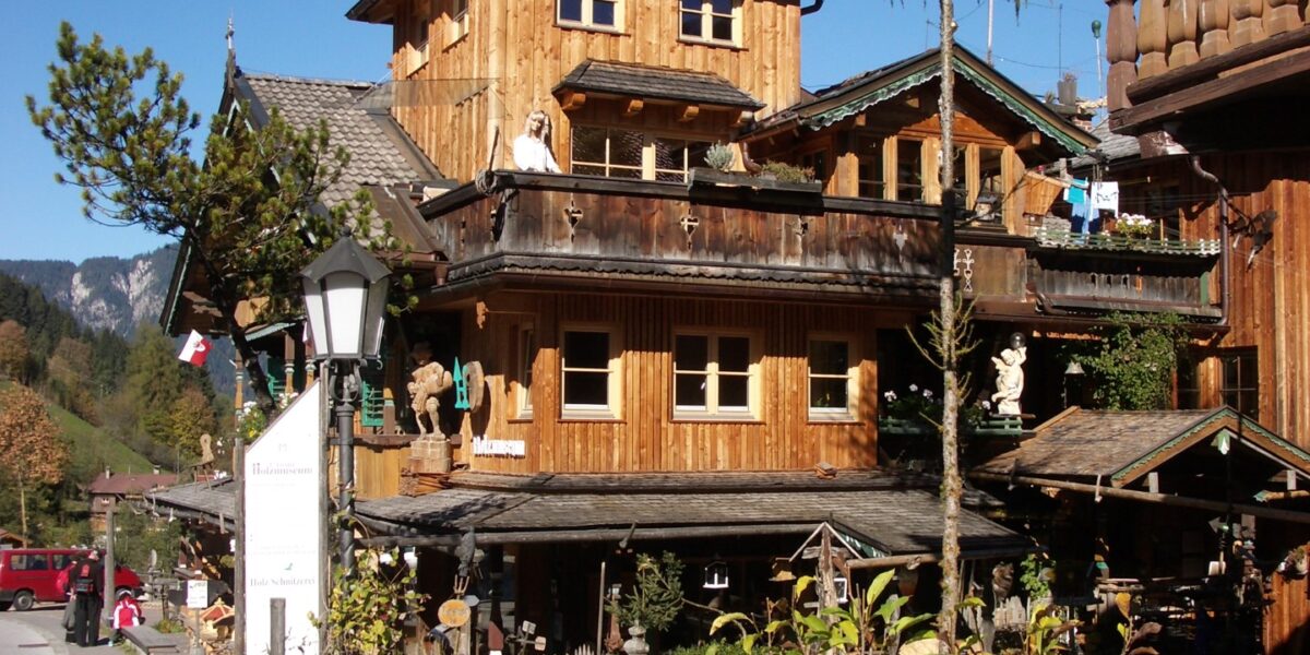 Holzhaus im Chalet-Stil mit Balkonen und geschnitzten Verzierungen, an einer Straße mit Bäumen und Bergen im Hintergrund unter blauem Himmel gelegen.