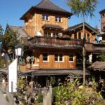 Holzhaus im Chalet-Stil mit Balkonen und geschnitzten Verzierungen, an einer Straße mit Bäumen und Bergen im Hintergrund unter blauem Himmel gelegen.