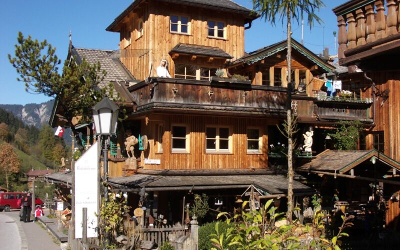 Holzhaus im Chalet-Stil mit Balkonen und geschnitzten Verzierungen, an einer Straße mit Bäumen und Bergen im Hintergrund unter blauem Himmel gelegen.