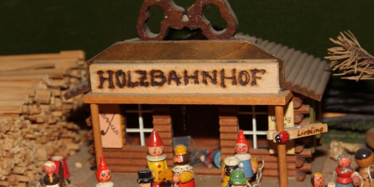 Holzspielzeugfiguren stehen vor einem kleinen Modellgebäude mit der Aufschrift "HOLZBAHNHOF", das von Holzstapeln und kleinen Schildern umgeben ist.