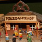 Holzspielzeugfiguren stehen vor einem kleinen Modellgebäude mit der Aufschrift "HOLZBAHNHOF", das von Holzstapeln und kleinen Schildern umgeben ist.