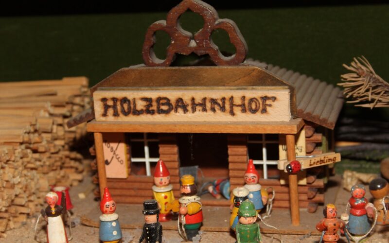 Holzspielzeugfiguren stehen vor einem kleinen Modellgebäude mit der Aufschrift "HOLZBAHNHOF", das von Holzstapeln und kleinen Schildern umgeben ist.
