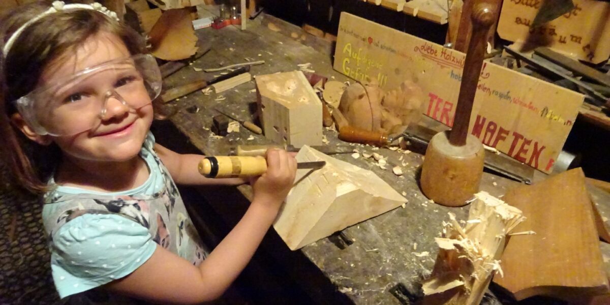 Ein junges Mädchen mit Schutzbrille arbeitet mit Meißel und Hammer an einer unübersichtlichen Werkbank in einer Holzwerkstatt, um Holz zu bearbeiten.