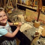 Ein junges Mädchen mit Schutzbrille arbeitet mit Meißel und Hammer an einer unübersichtlichen Werkbank in einer Holzwerkstatt, um Holz zu bearbeiten.