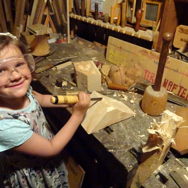 Ein junges Mädchen mit Schutzbrille arbeitet mit Meißel und Hammer an einer unübersichtlichen Werkbank in einer Holzwerkstatt, um Holz zu bearbeiten.