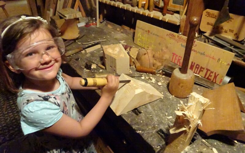 Ein junges Mädchen mit Schutzbrille arbeitet mit Meißel und Hammer an einer unübersichtlichen Werkbank in einer Holzwerkstatt, um Holz zu bearbeiten.