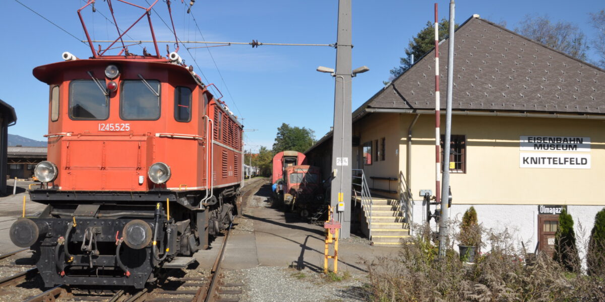 Rote historische Elektrolokomotive auf den Gleisen neben dem Gebäude des Eisenbahnmuseums Knittelfeld, mit einem weiteren Zug und einem Museumsschild im Hintergrund.