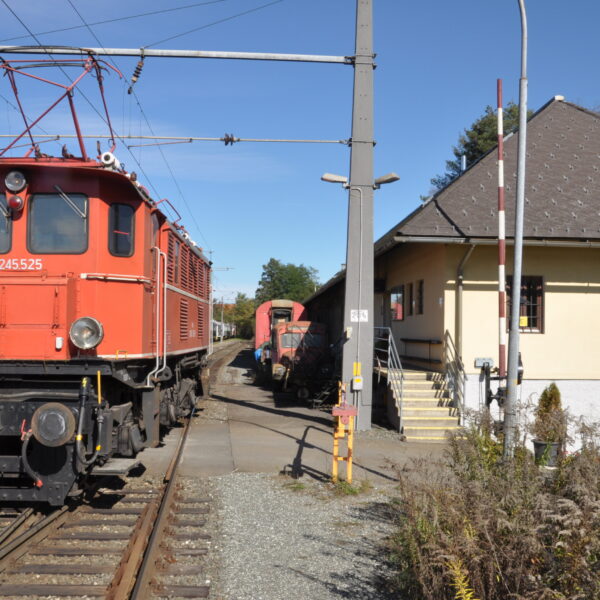 Rote historische Elektrolokomotive auf den Gleisen neben dem Gebäude des Eisenbahnmuseums Knittelfeld, mit einem weiteren Zug und einem Museumsschild im Hintergrund.