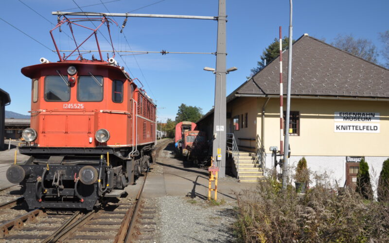 Rote historische Elektrolokomotive auf den Gleisen neben dem Gebäude des Eisenbahnmuseums Knittelfeld, mit einem weiteren Zug und einem Museumsschild im Hintergrund.