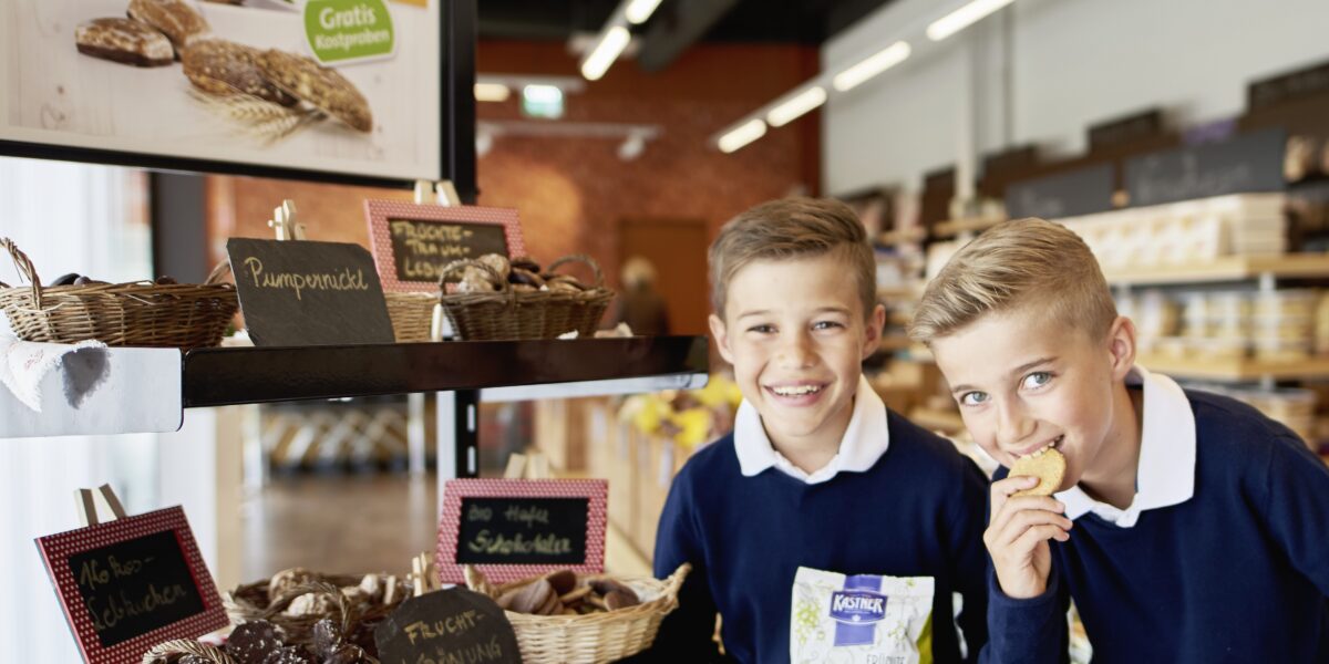 Zwei Jungen in blauen Pullovern stehen vor einer Bäckerei mit verschiedenen Brotsorten und Gebäck; einer isst einen Keks, der andere lächelt und hält eine Tüte mit Keksen in der Hand.