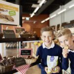 Zwei Jungen in blauen Pullovern stehen vor einer Bäckerei mit verschiedenen Brotsorten und Gebäck; einer isst einen Keks, der andere lächelt und hält eine Tüte mit Keksen in der Hand.