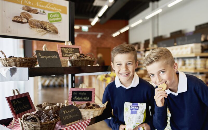 Zwei Jungen in blauen Pullovern stehen vor einer Bäckerei mit verschiedenen Brotsorten und Gebäck; einer isst einen Keks, der andere lächelt und hält eine Tüte mit Keksen in der Hand.