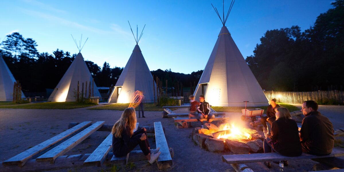 Menschen sitzen in der Abenddämmerung auf Holzbänken um ein Lagerfeuer herum, im Hintergrund stehen mehrere große Tipis, die von Lichtern beleuchtet werden.