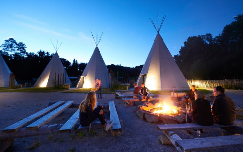 Menschen sitzen in der Abenddämmerung auf Holzbänken um ein Lagerfeuer herum, im Hintergrund stehen mehrere große Tipis, die von Lichtern beleuchtet werden.