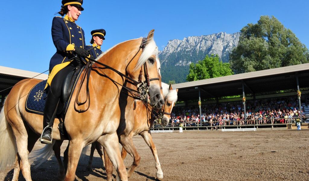 Zwei uniformierte Reiter auf braunen Pferden treten in einer Freiluftarena vor Publikum auf, im Hintergrund sind ein Berg und Bäume zu sehen.