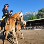 Zwei uniformierte Reiter auf braunen Pferden treten in einer Freiluftarena vor Publikum auf, im Hintergrund sind ein Berg und Bäume zu sehen.
