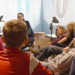 Eine Gruppe von Kindern sitzt in einem Kreis auf dem Boden und hört einem Erwachsenen zu, der in einem hell erleuchteten Raum vor ihnen spricht.