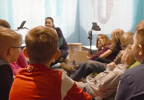 Eine Gruppe von Kindern sitzt in einem Kreis auf dem Boden und hört einem Erwachsenen zu, der in einem hell erleuchteten Raum vor ihnen spricht.