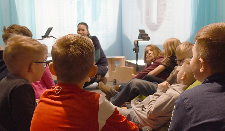 Eine Gruppe von Kindern sitzt in einem Kreis auf dem Boden und hört einem Erwachsenen zu, der in einem hell erleuchteten Raum vor ihnen spricht.