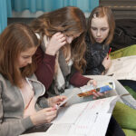 Drei Mädchen im Teenageralter sitzen auf dem Boden, sehen sich Papiere und Zeitschriften an und scheinen gemeinsam an einem Projekt oder einer Aufgabe zu arbeiten.