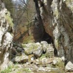 Eine kleine Holzleiter lehnt an Felsen in einer engen, felsigen Bergschlucht, durch die ein Bach fließt. Im Hintergrund sind spärliche Bäume zu sehen.