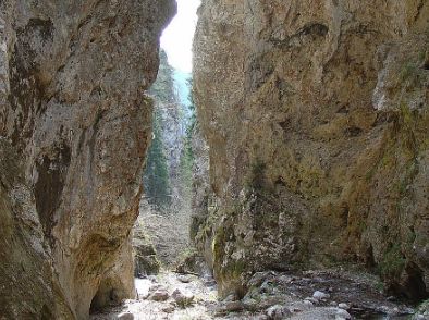 Enge, felsige Schlucht mit steilen Klippen auf beiden Seiten, etwas Grün in der Ferne und einem felsigen Bachbett am Fuße.