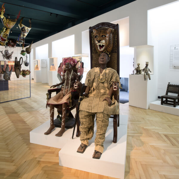 Eine Museumsausstellung zeigt afrikanische Stühle, Masken und Skulpturen, darunter zwei Schaufensterpuppen in traditioneller Kleidung, die auf kunstvollen Thronen sitzen.