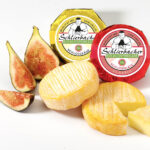 Zwei ganze und eine in Scheiben geschnittene Feigen sind neben zwei runden Stücken Schlierbacher Käse ausgestellt, von denen eines teilweise aufgeschnitten ist, mit einer beschrifteten Verpackung im Hintergrund.