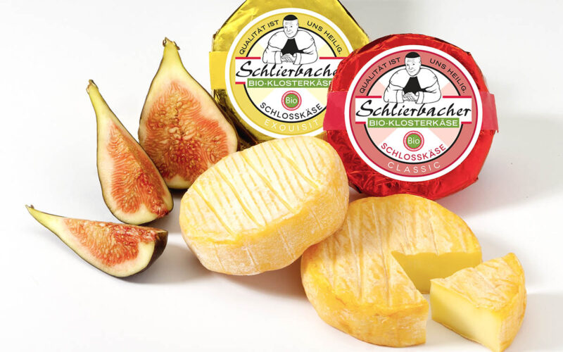 Zwei ganze und eine in Scheiben geschnittene Feigen sind neben zwei runden Stücken Schlierbacher Käse ausgestellt, von denen eines teilweise aufgeschnitten ist, mit einer beschrifteten Verpackung im Hintergrund.