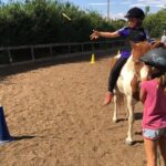 Ein Kind auf einem Pferd wirft einen Ring in Richtung eines Kegels, während ein anderes Kind mit Helm in der Nähe eines Reitplatzes steht.