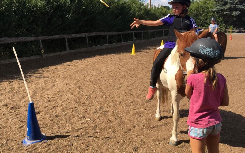 Ein Kind auf einem Pferd wirft einen Ring in Richtung eines Kegels, während ein anderes Kind mit Helm in der Nähe eines Reitplatzes steht.