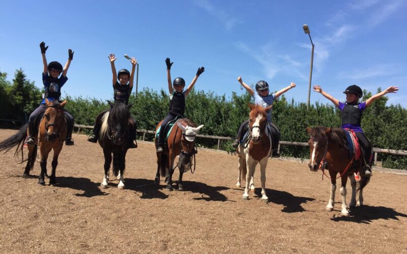 Fünf Kinder mit Helmen sitzen in einer Reihe auf Pferden und heben ihre Arme in die Luft, auf einem unbefestigten Reitplatz mit Bäumen und einem blauen Himmel im Hintergrund.