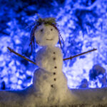 Ein kleiner Schneemann mit Zweigarmen, einem lächelnden Gesicht und Knopfdetails steht nachts draußen, umgeben von Schnee und von blauen Lichtern beleuchtet.