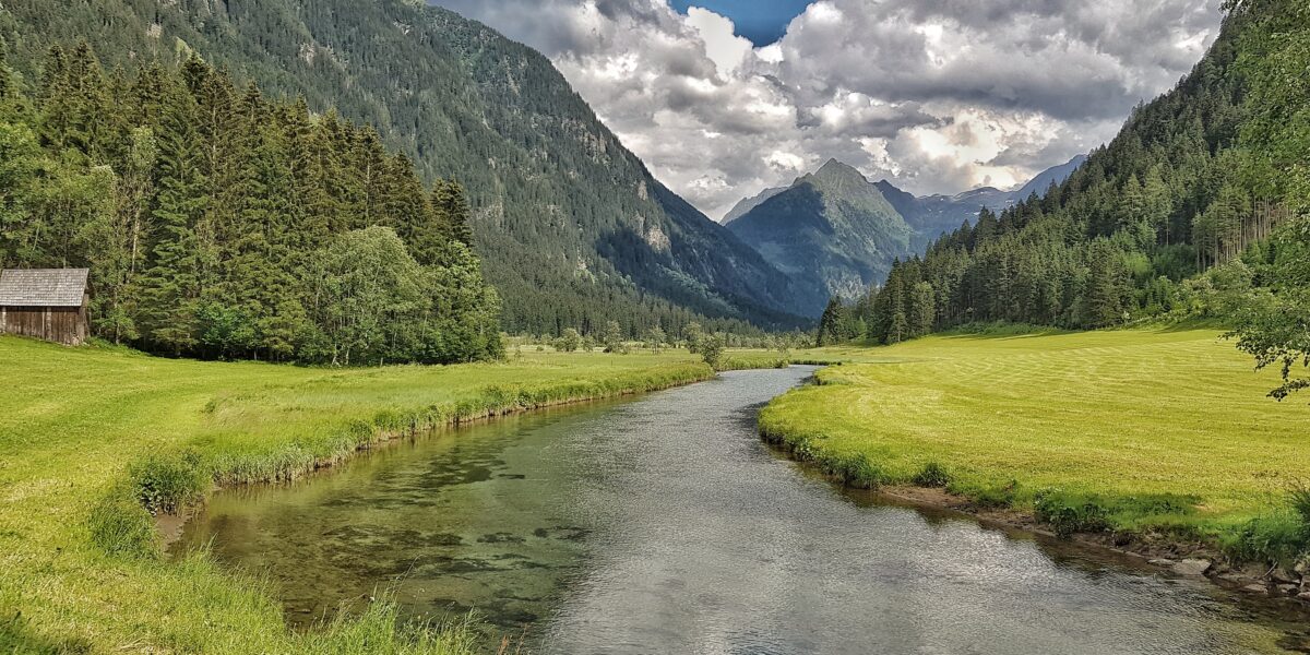 Ein klarer Fluss fließt durch ein grünes Tal, das von dichten Wäldern und hohen Bergen unter einem teilweise bewölkten Himmel umgeben ist.