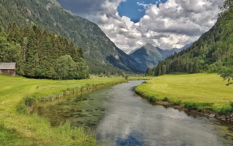 Ein klarer Fluss fließt durch ein grünes Tal, das von dichten Wäldern und hohen Bergen unter einem teilweise bewölkten Himmel umgeben ist.