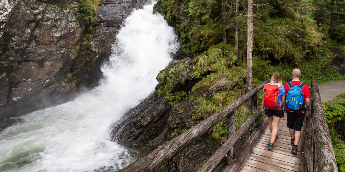 Zwei Wanderer mit Rucksäcken überqueren eine Holzbrücke neben einem rauschenden Wasserfall in einer bewaldeten, felsigen Landschaft.