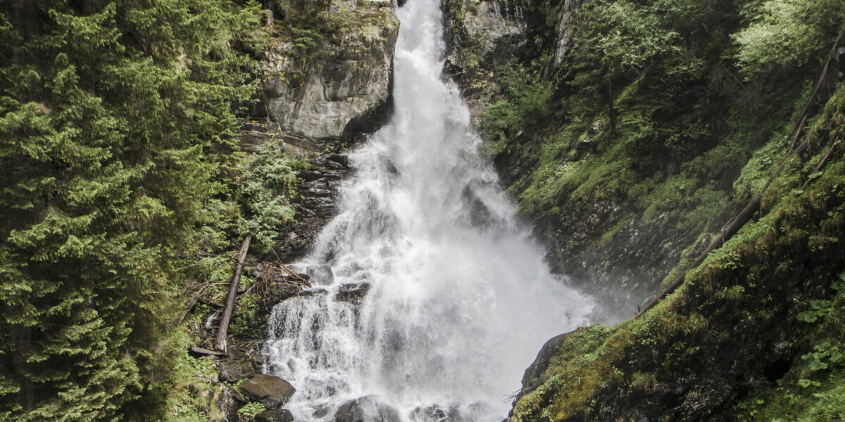 Ein hoher Wasserfall stürzt in Kaskaden eine von dichten Kiefern umgebene Felswand hinunter, über die eine kleine Brücke führt, und der Himmel ist teilweise bewölkt.