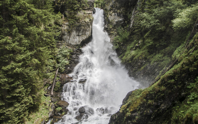 Ein hoher Wasserfall stürzt in Kaskaden eine von dichten Kiefern umgebene Felswand hinunter, über die eine kleine Brücke führt, und der Himmel ist teilweise bewölkt.