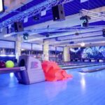 Modernes Interieur einer Bowlingbahn mit mehreren Bahnen, bunten Bowlingkugeln auf einem Gestell, orangefarbenen Kunststofframpen und stimmungsvoller Beleuchtung.