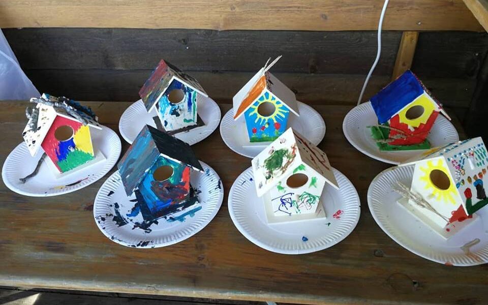 Sechs bemalte hölzerne Vogelhäuschen stehen auf Papptellern auf einem Holztisch, jedes mit verschiedenen bunten Motiven verziert.