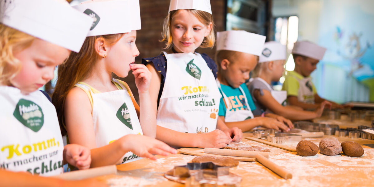 Mehrere Kinder mit Schürzen und Kochmützen stehen an einem bemehlten Tisch und stellen in einer hellen Küchenumgebung mit Nudelholz und Ausstechern Teig her.