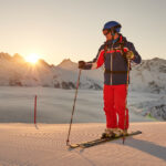 Ein Skifahrer in roter und blauer Ausrüstung steht bei Sonnenaufgang auf einer präparierten Piste, im Hintergrund schneebedeckte Berge und eine Nebelschicht.