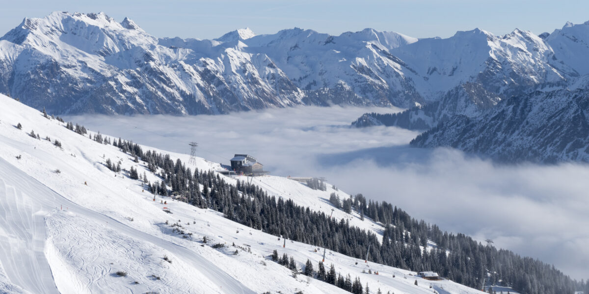 Schneebedeckte Berge mit Skipisten, immergrünen Bäumen und einer Hütte; tief hängende Wolken füllen das Tal, und im Hintergrund sind bei klarem Himmel schroffe Gipfel zu sehen.